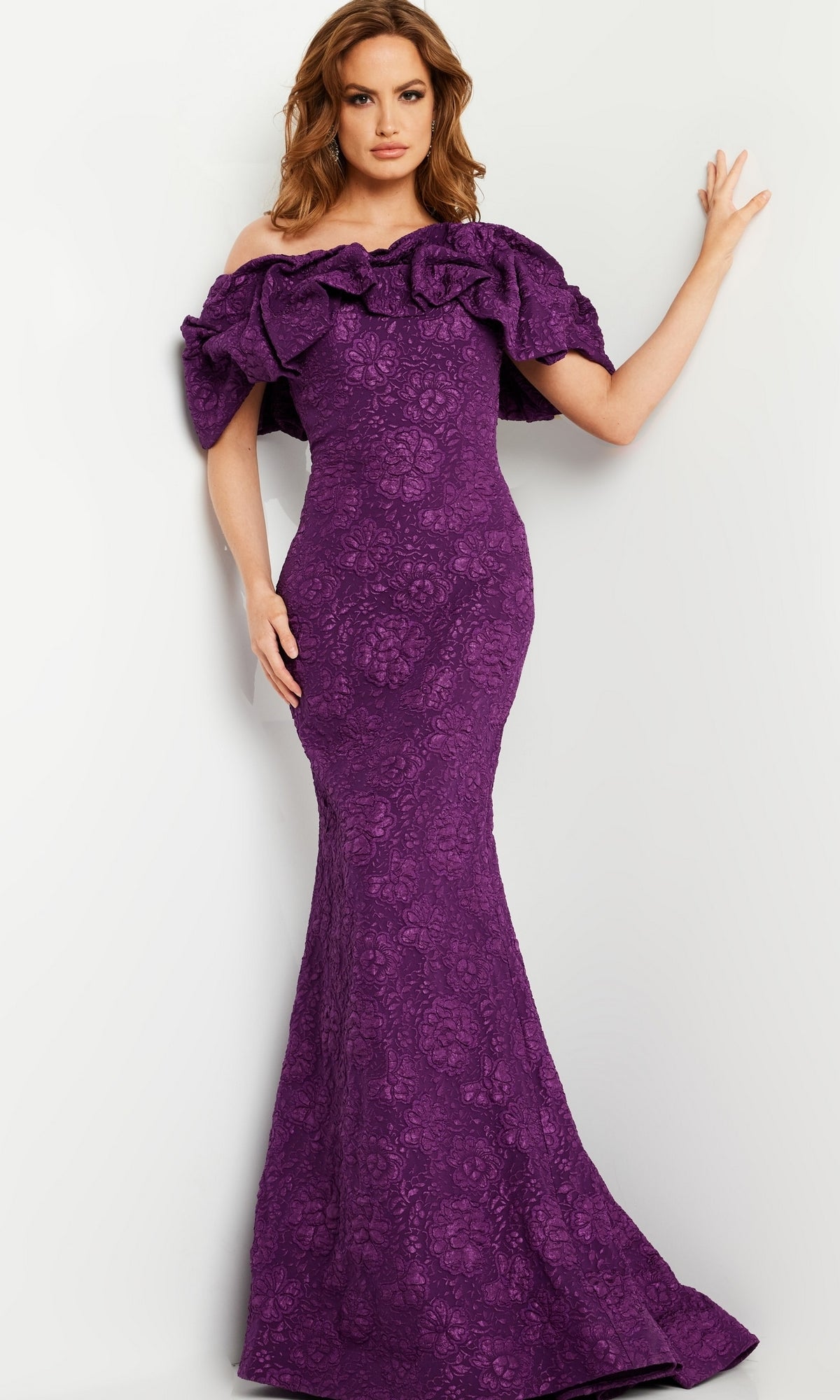 Woman wearing the Jovani Long Brocade Mermaid Prom Dress 23847, a fitted, floor-length purple gown with an off-the-shoulder ruffled neckline, standing against a plain light-colored wall.